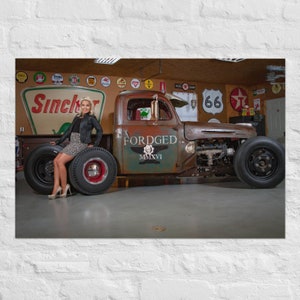 Könnte beinhalten: Ein rostiger brauner Vintage-Pickup mit einer Frau, die auf dem Hinterrad sitzt. Der Truck hat eine individuelle Lackierung mit den Worten "FORDGED MMXVI" an der Seite. Der Truck steht in einer Garage mit verschiedenen Schildern und Dekorationen an den Wänden.