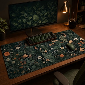 Op de afbeelding: Een rechthoekige bureaumat met een donkergroene achtergrond en een dicht botanisch patroon van diverse bloemen en bladeren in tinten oranje, wit en gedempt groen. De mat ligt op een houten bureau onder een computermonitor.