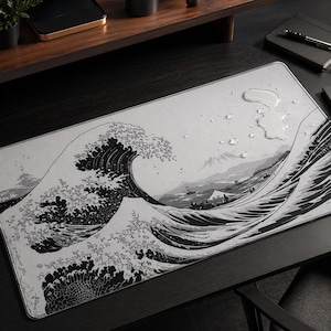 May include: A rectangular desk mat featuring a black and white graphic print of a large ocean wave, similar to the Great Wave off Kanagawa. The surface of the mat is light gray with dark ink-style illustrations of water, boats, and a mountain.