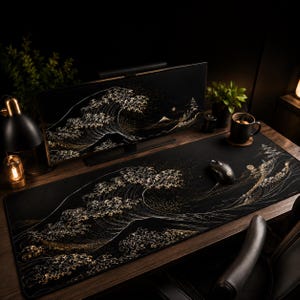 May include: A large rectangular desk mat featuring a black background with a gold and white stylized wave design. The mat sits on a wooden desk surface alongside a black computer mouse, a black mug, and a computer monitor displaying the same wave art.