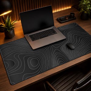 May include: A large rectangular desk mat featuring a black surface with thin, white, flowing topographic contour lines. The mat is positioned on a wooden desk surface underneath a silver laptop and a black computer mouse.