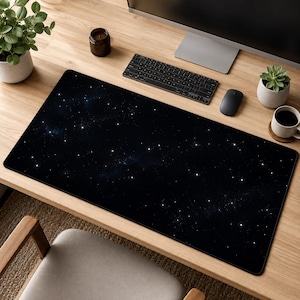 以下が含まれることがあります： 散りばめられた白い星と淡い星雲が描かれた、深い黒色の宇宙柄の長方形デスクマット。マットは滑らかな表面で、木製のデスクの上に配置されており、コンピューター周辺機器のための広い作業スペースを提供しています。