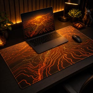 May include: A large rectangular desk mat featuring a dark background with glowing orange topographic contour lines. The mat sits under a laptop and a black computer mouse, displaying a continuous pattern of flowing, abstract orange lines.