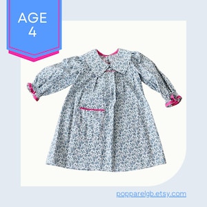 May include: A white dress with a blue floral print and pink trim. The dress has a Peter Pan collar, long sleeves, and a single pocket on the front. The dress is for a child and has the text "AGE 4" on the label.