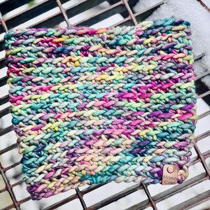 May include: A hand-knitted cowl in a vibrant mix of colors including pink, purple, teal, and yellow. The chunky knit scarf is made of thick yarn and features a small leather tag with the text "Mama Cowl". The cowl is rectangular in shape.