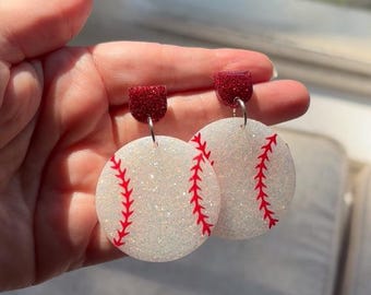 May include: A pair of baseball-themed earrings. Each earring features a white, glittery, circular design with red stitching, mimicking a baseball. The top portion of each earring is a small, rectangular, glittery red stud.