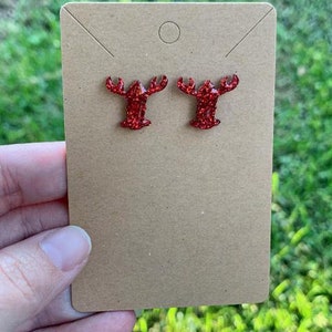 May include: A pair of red glitter lobster earrings. The earrings are on a brown card.