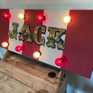 Puede incluir: Un letrero rectangular de madera con el nombre "JACK" en una fuente de circo vintage. El letrero tiene rayas rojas y blancas alternas y está iluminado por una serie de bombillas rojas y blancas.