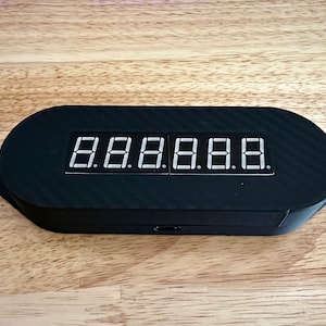 May include: Black electronic device with a digital display showing the numbers 8.8.8.8.8.8. The device has a carbon fiber texture and an oval shape. The display is white and the device is on a wooden surface.