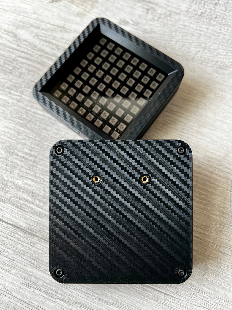 May include: A black square box with a carbon fiber pattern and two gold screws. The box has a small square opening on the top with a grid of small black squares.