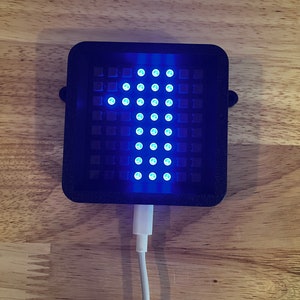 May include: A black rectangular device with a grid of blue lights forming an arrow pointing upwards. The device is plugged into a white USB cable.