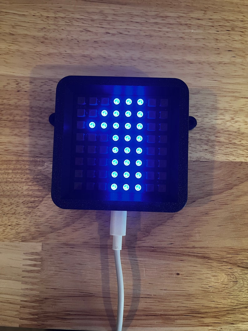 May include: A black rectangular device with a grid of blue lights forming an arrow pointing upwards. The device is plugged into a white USB cable.