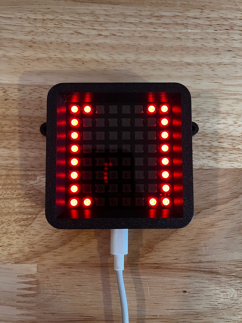 May include: A black rectangular device with a grid of red LED lights. The device is plugged into a white USB cable.