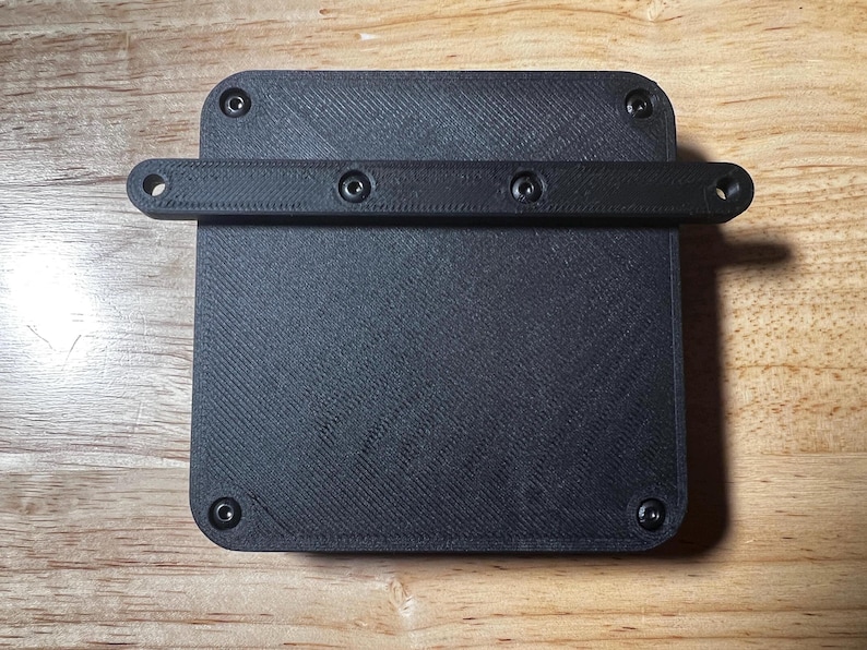 May include: Black plastic box with a textured surface and four screw holes. A black plastic bar with three screw holes is attached to the top of the box.