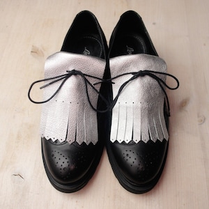 May include: A pair of black leather shoes with silver fringe detailing. The shoes have a lace-up closure and a rounded toe.
