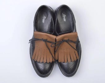 May include: A pair of black leather shoes with brown leather fringe accents. The shoes have a classic design with decorative perforations and a leather tongue. The shoes are displayed on a white background.