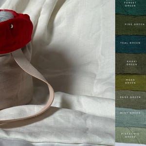 May include: A beige linen tote bag with a red fabric lining and two cream-colored handles. The bag is filled with yarn balls in various shades of blue and gray. The image also shows a color chart with swatches of different shades of green and red.