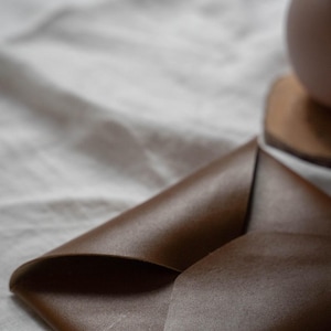 Puede incluir: Una cartera de cuero marr&oacute;n de estilo sobre, doblada y cerrada. La cartera est&aacute; sobre una superficie de tela blanca.