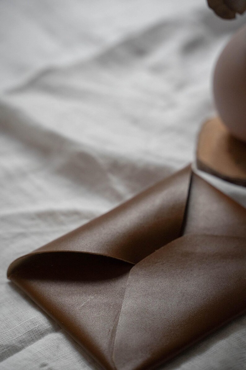 Puede incluir: Una cartera de cuero marr&oacute;n de estilo sobre, doblada y cerrada. La cartera est&aacute; sobre una superficie de tela blanca.