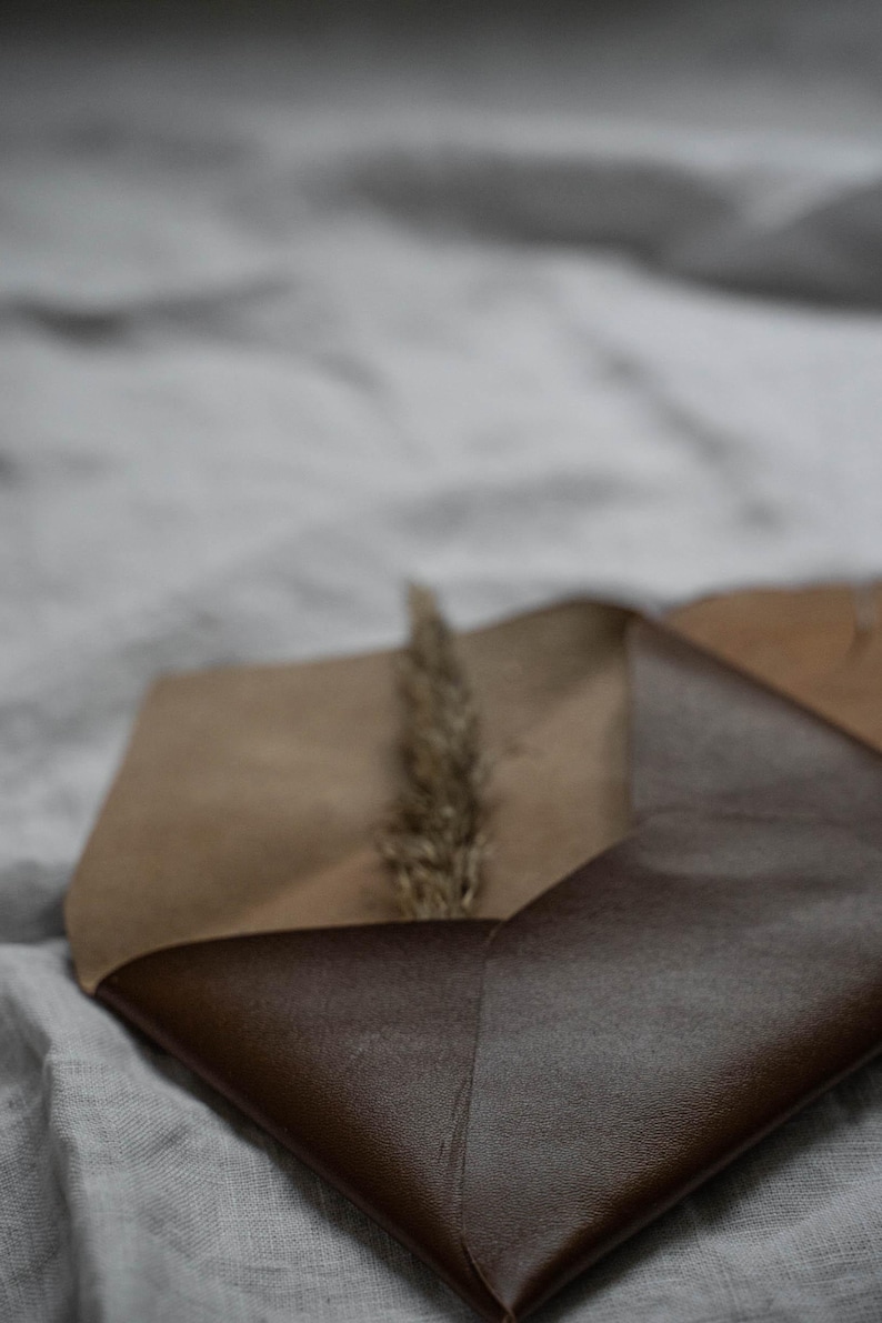 Puede incluir: Un sobre de cuero marr&oacute;n con una solapa cerrada sobre un trozo de papel marr&oacute;n. Un tallo de hierba seca es visible dentro del sobre.