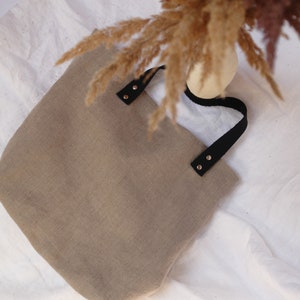 Minimalist linen tote bag with black leather handles and dried pampas grass, set against a neutral fabric backdrop.