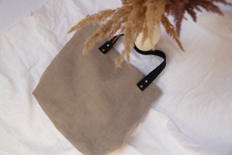Minimalist linen tote bag with black leather handles and dried pampas grass, set against a neutral fabric backdrop.