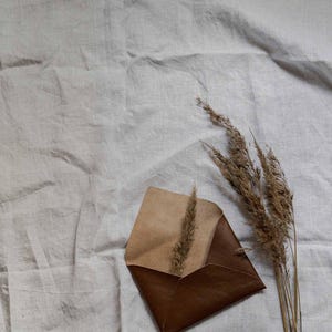 Puede incluir: Un sobre de cuero marr&oacute;n con una solapa beige, sobre un fondo de tela de lino blanco. Un manojo de hierba seca est&aacute; a la derecha del sobre.