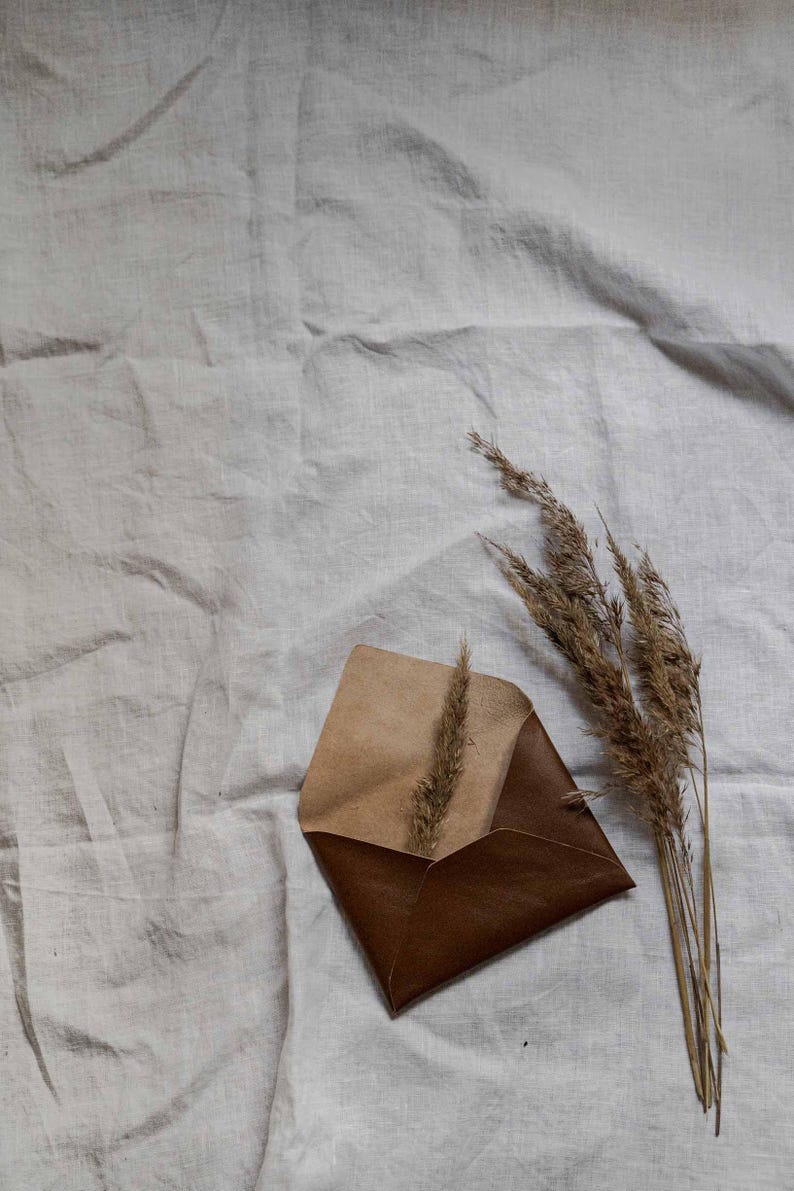 Puede incluir: Un sobre de cuero marr&oacute;n con una solapa beige, sobre un fondo de tela de lino blanco. Un manojo de hierba seca est&aacute; a la derecha del sobre.
