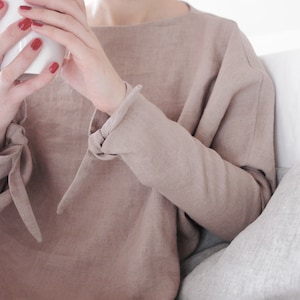 Minimalist linen kimono-style top in a neutral beige tone. Features long sleeves with delicate tie details at the wrists and a relaxed, oversized fit. Made from breathable, high-quality linen for effortless comfort and style