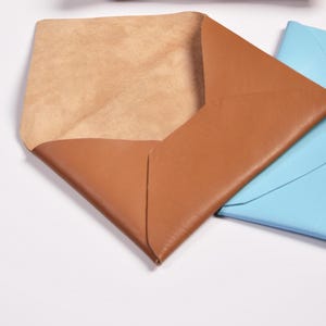 Puede incluir: Tres sobres de cuero en diferentes colores: marr&oacute;n, azul y beige. El sobre marr&oacute;n est&aacute; abierto, revelando un interior beige.