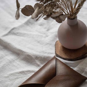 Puede incluir: Un sobre de cuero marr&oacute;n sobre un mantel de lino blanco junto a un jarr&oacute;n con flores secas y un posavasos de madera.