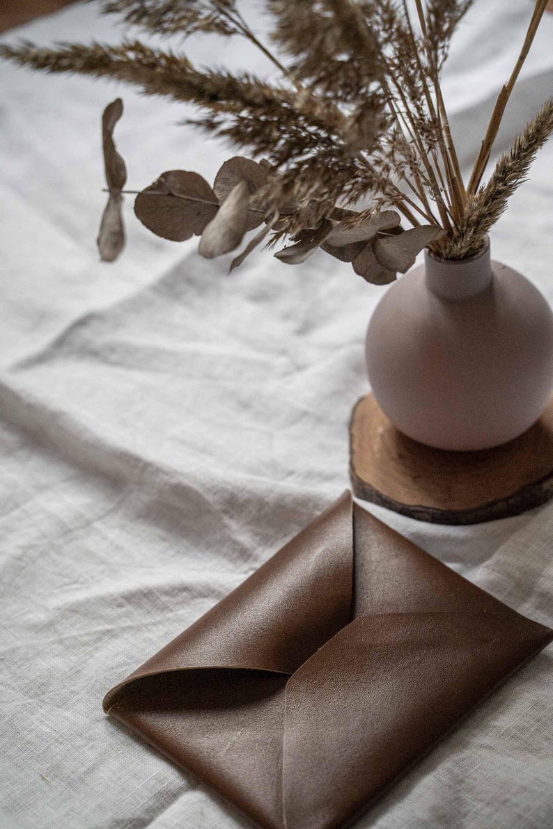 Puede incluir: Un sobre de cuero marr&oacute;n sobre un mantel de lino blanco junto a un jarr&oacute;n con flores secas y un posavasos de madera.