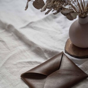 Puede incluir: Un sobre de cuero marr&oacute;n sobre una superficie de lino blanco. El sobre est&aacute; doblado por la mitad y tiene un brillo sutil.