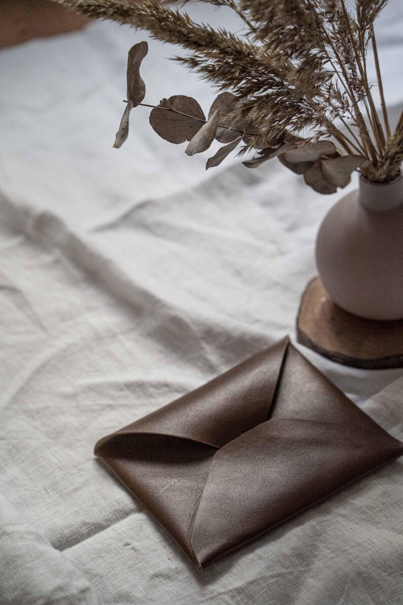 Puede incluir: Un sobre de cuero marr&oacute;n sobre una superficie de lino blanco. El sobre est&aacute; doblado por la mitad y tiene un brillo sutil.