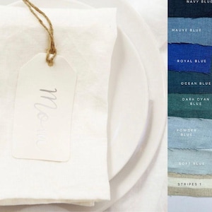 May include: A white linen napkin with a white tag that says "Mon" on it. The napkin is on a white plate. The image is a close-up of the napkin and tag.