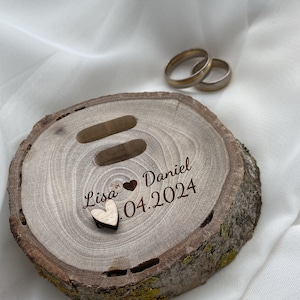 Könnte beinhalten: Holzringkissen mit zwei Schlitzen für Ringe. Die Box ist mit den Namen "Lisa" und "Daniel" sowie dem Datum "04.2024" graviert. Ein kleines Holzhäuschen befindet sich auf der Box.