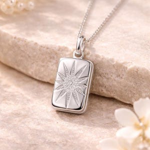 Op de afbeelding: Zilveren ketting met rechthoekige hanger met een sterrenpatroon. Het sterrenpatroon is ingelegd met kleine, sprankelende steentjes. De ketting wordt getoond op een lichtgekleurd, getextureerd oppervlak.
