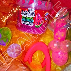 Op de afbeelding: Een verzameling kleurrijk speelgoed en slijm. De afbeelding toont een container met "Crazy Aaron's Slime Charmers" met watermeloenafbeeldingen, een roze bellenstaf, een roze en oranje vorm en een groen en paars hartvormig pop-it speelgoed.