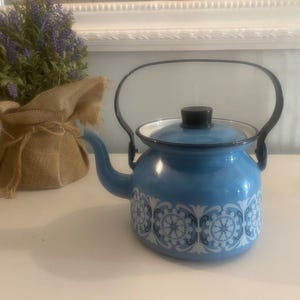 Puede incluir: Una tetera de esmalte azul claro con asa y tapa negras. La tetera presenta un estampado floral blanco. Una pequeña bolsa de arpillera con flores moradas está en el fondo. Aproximadamente 20 cm de altura.