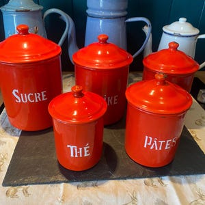 Könnte beinhalten: Eine Sammlung roter Emaille-Küchenbehälter mit weißer Schrift, darunter "SUCRE", "FARINE", "THÉ" und "PÂTES". Die Behälter haben passende Deckel und stehen auf einer schwarzen Schieferplatte. Im Hintergrund sind mehrere Vintage-Teekannen zu sehen.