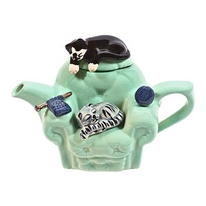 以下が含まれることがあります： 蓋に黒い猫の置物、椅子に座っているグレーと白の猫の置物がついた、アームチェアの形をしたターコイズブルーのセラミックティーポット。ティーポットには、青い毛糸玉と編み針が側面についています。