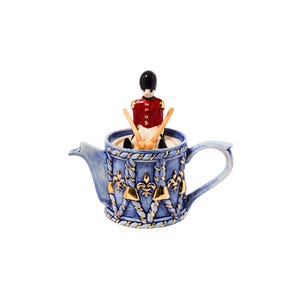 May include: A blue ceramic teapot with gold detailing and a figurine of a soldier inside. The teapot has a handle and spout, with a rope and fleur-de-lis pattern. The soldier wears a red jacket and a black hat.