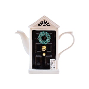May include: A novelty teapot in the shape of a front door. The black door features a green wreath and gold door furniture. The white teapot has a spout, handle, and a small sign that says "TEA TIMES".