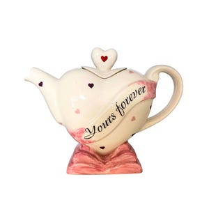 Könnte beinhalten: Weiße Keramik-Teekanne in Herzform mit einem rosa Band, auf dem "Yours forever" steht. Die Teekanne hat einen herzförmigen Deckel mit einem roten Herzen oben drauf. Die Teekanne ist mit kleinen rosa und violetten Herzen verziert.