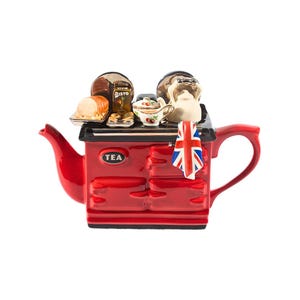 Può includere: Teiera in ceramica rossa a forma di stufa, con manico e beccuccio. La teiera ha un canovaccio Union Jack, una piccola teiera e generi alimentari sopra. La parola "TEA" è stampata sul davanti.