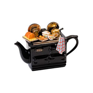 Könnte beinhalten: Eine schwarze Keramik-Teekanne in Form eines Ofens, mit Griff und Ausguss. Auf der Oberseite des Ofens befinden sich Miniatur-Lebensmittel, darunter ein Brotlaib, ein Bisto-Behälter und eine kleine Teetasse. Ein kariertes Geschirrtuch hängt an der Seite. Das Wort "TEA" steht auf der Vorderseite.