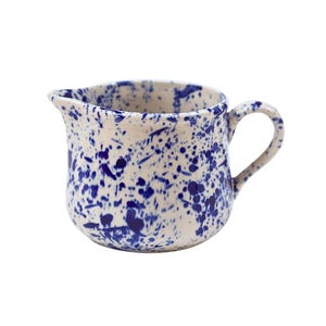 May include: A white ceramic creamer with a blue splatter design. The creamer has a rounded body and a handle.