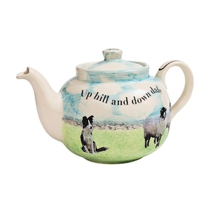 Puede incluir: Una tetera de cerámica blanca con un diseño azul y verde que presenta un border collie blanco y negro y dos ovejas. La tetera tiene un asa y un pico y dice "Up hill and down dale".