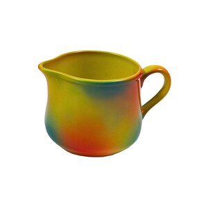 May include: A yellow, green, orange and red ceramic creamer with a rounded body and a handle.