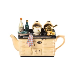 May include: A cream-coloured teapot shaped like a vintage stove with black accents. The teapot has a gold-coloured tea pot lid and a black tea kettle on top. The stove has a black oven door with the word "TEA" written on it. There are various kitchen accessories on top of the stove, including a small pot, a bottle, and a dish towel.